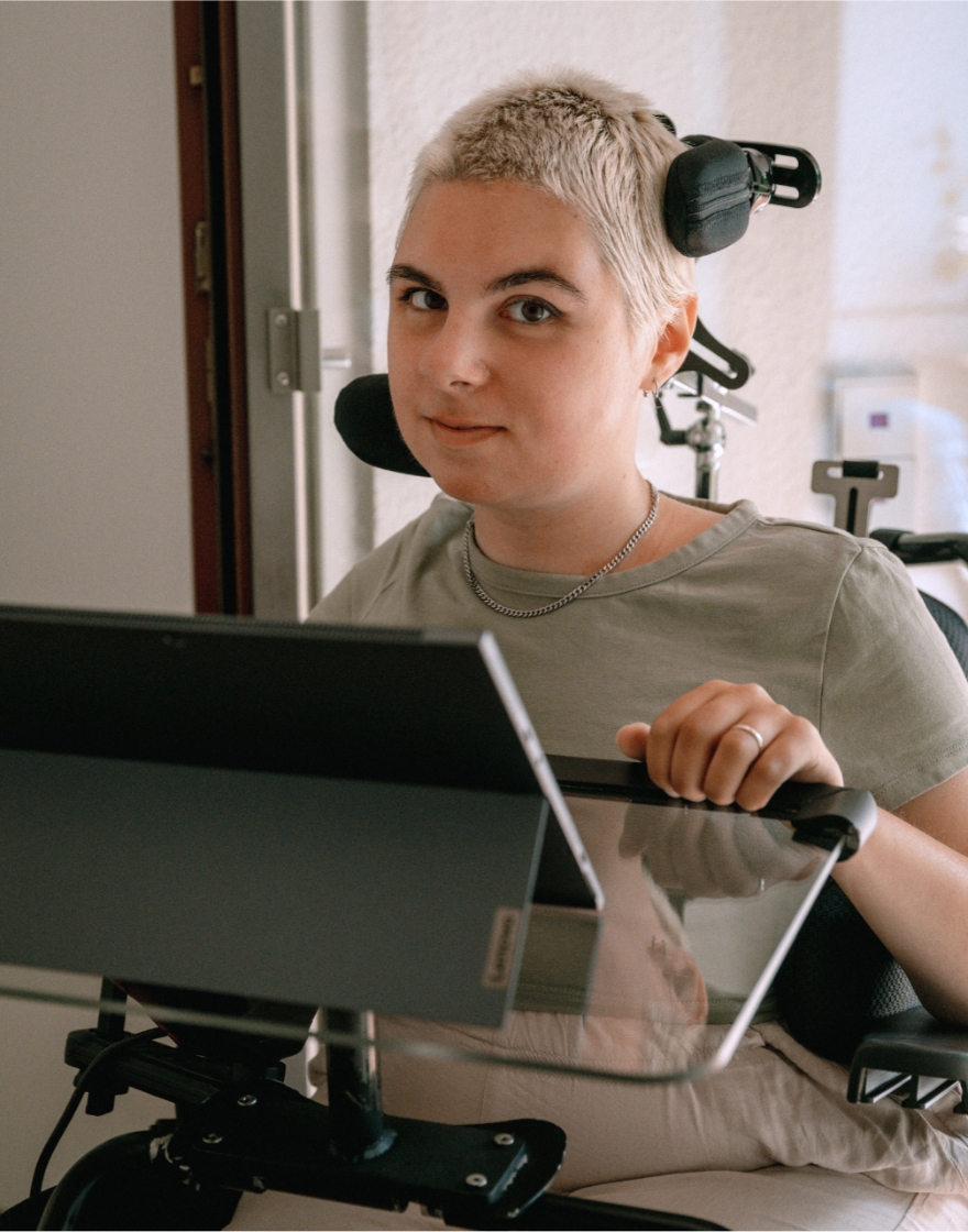 Eliza Gawin sitzt im Rollstuhl, hat blonde Haare, sitzt an einem Rollstuhltisch, darauf steht ein Laptop.